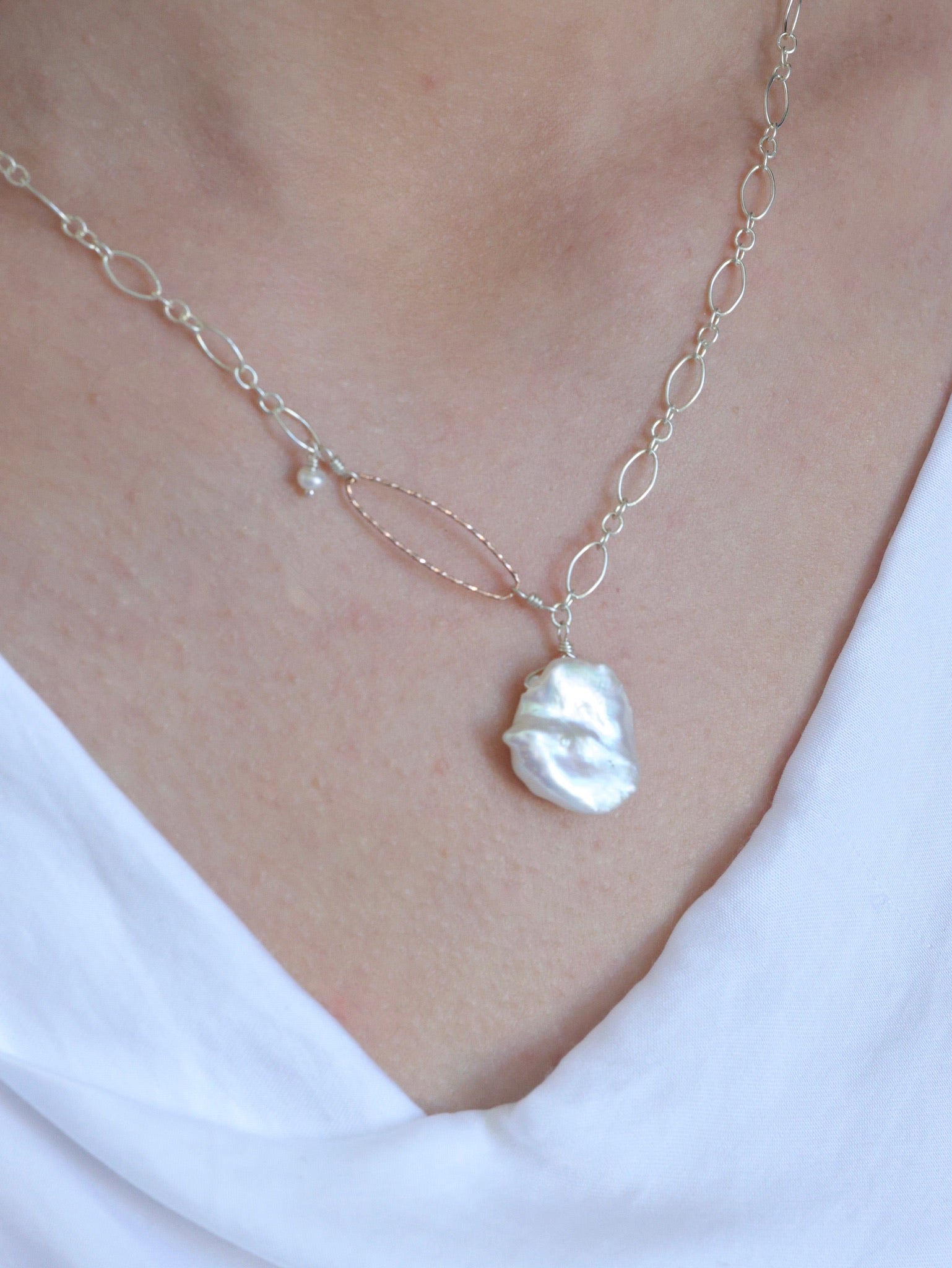 Woman wearing the Veronica White Keshi Freshwater Pearl Necklace, featuring a large petal-shaped pearl on a sterling silver chain with a hammered rose gold loop and small pearl detail. Shown styled with a white draped neckline top.