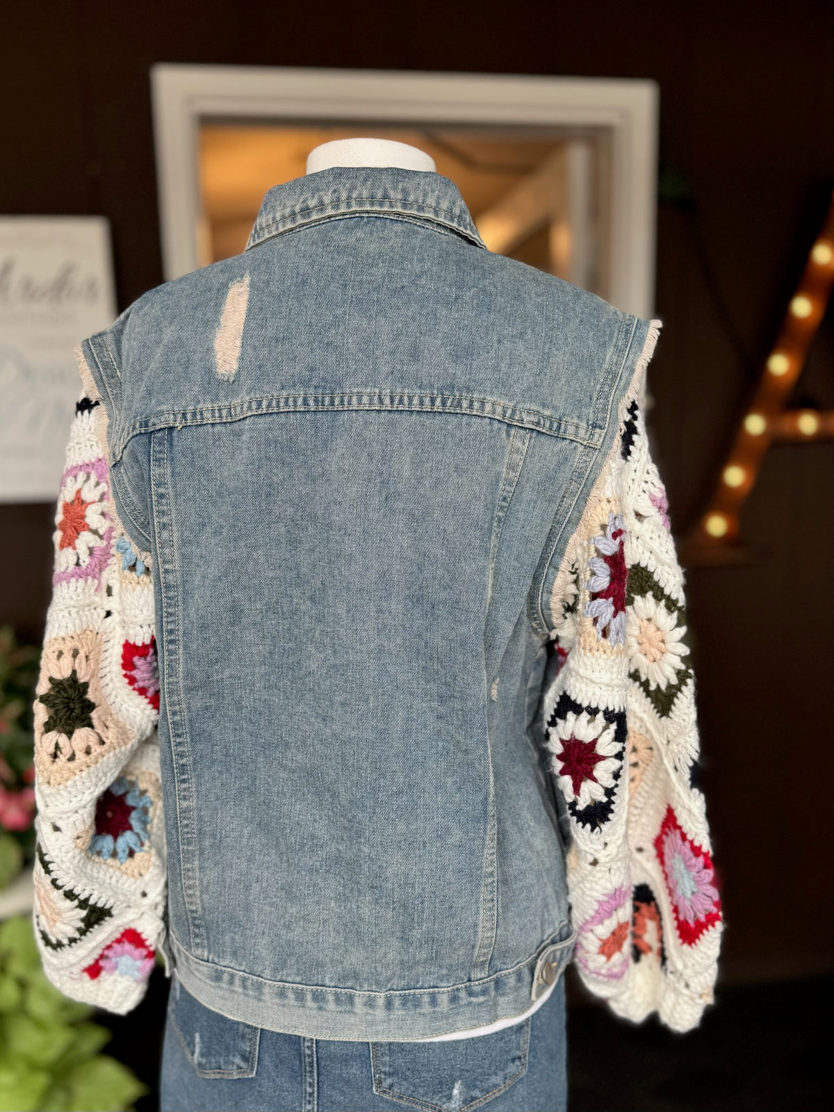 Back view of the Dahlia Granny Square Denim Jacket showing light wash denim with mild distressing and hand-crocheted Sunrise Ivory granny square sleeves at Ardor Boutique.
