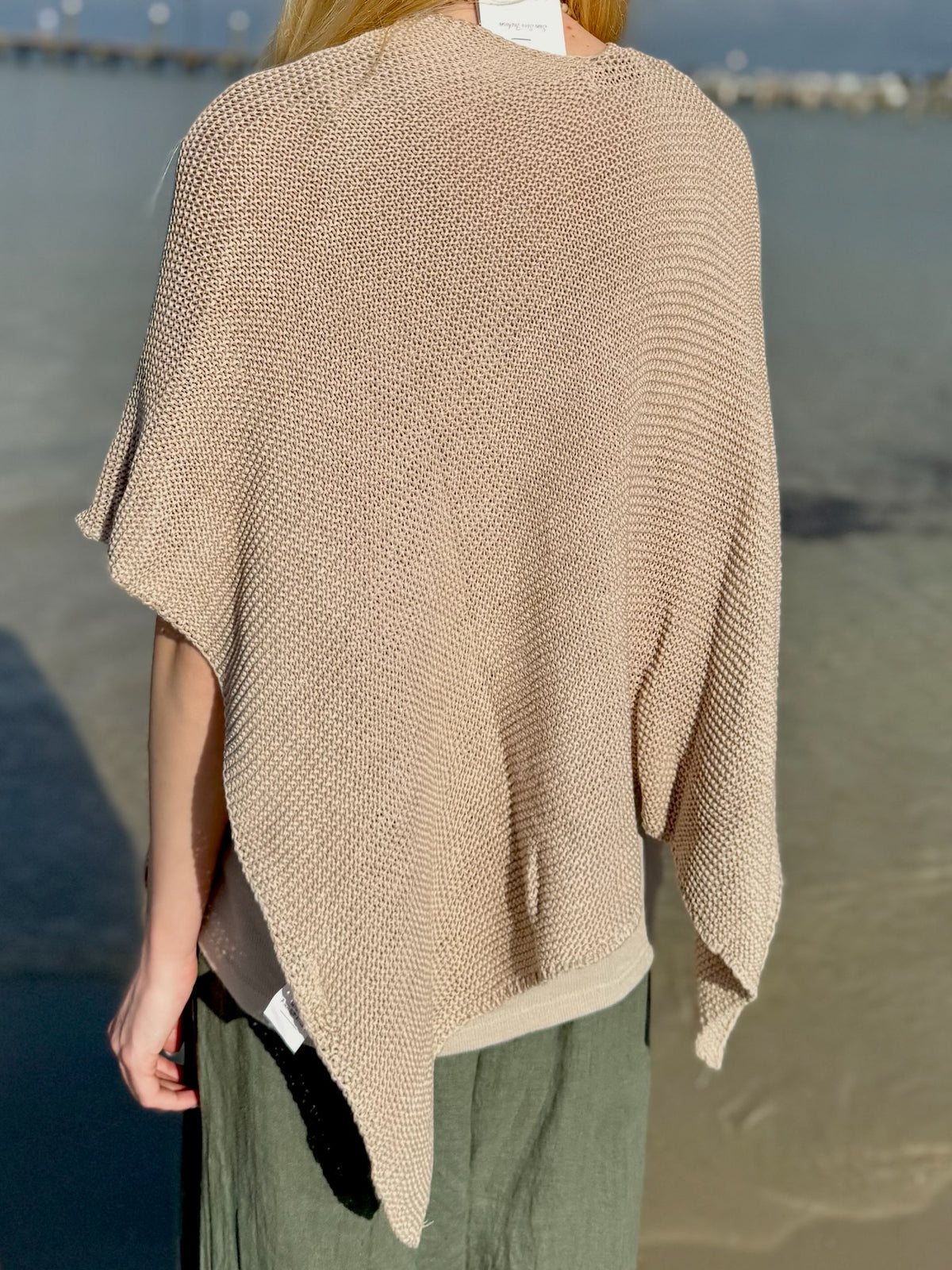 Back view of the Elise Wrap Poncho in Beige featuring a textured knit, relaxed drape, and asymmetrical hem, styled with olive green pants at Ardor Boutique.
