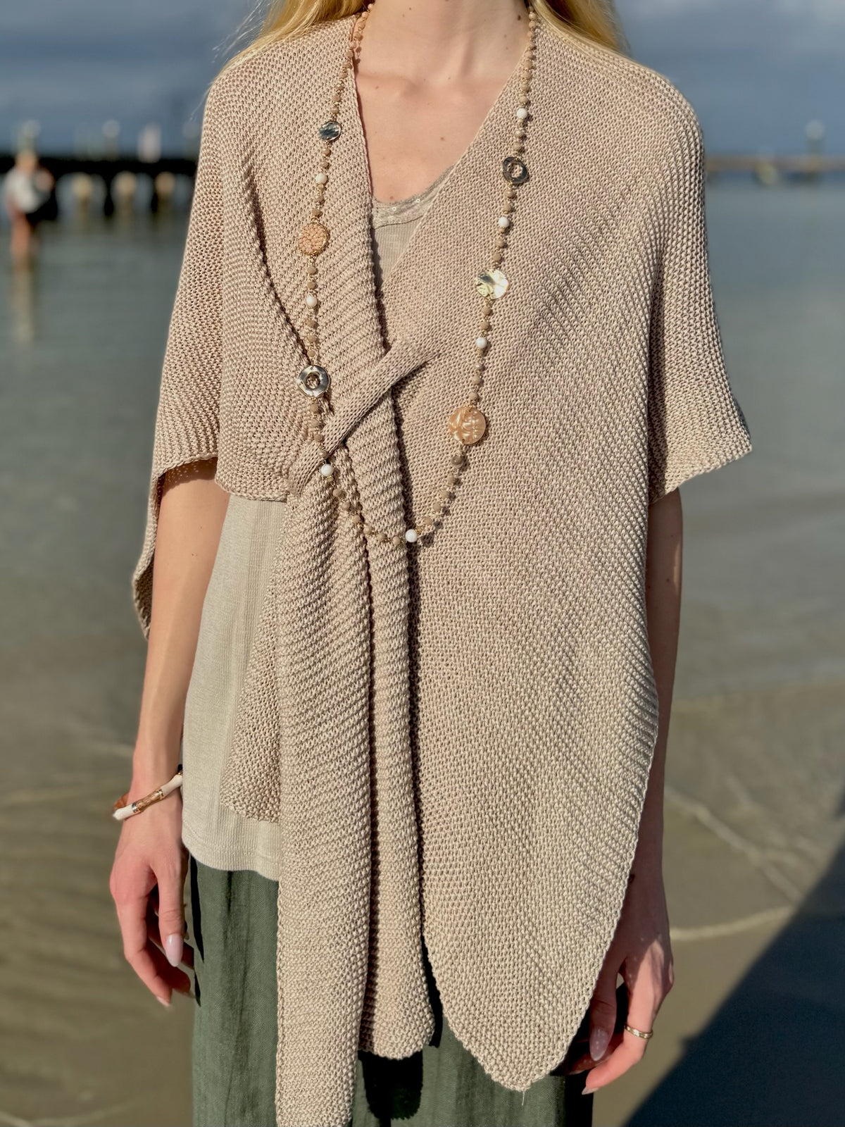 Front view of the Elise Wrap Poncho in Beige featuring a textured knit, soft draping, and front tie detail, styled with a long beaded necklace at Ardor Boutique.
