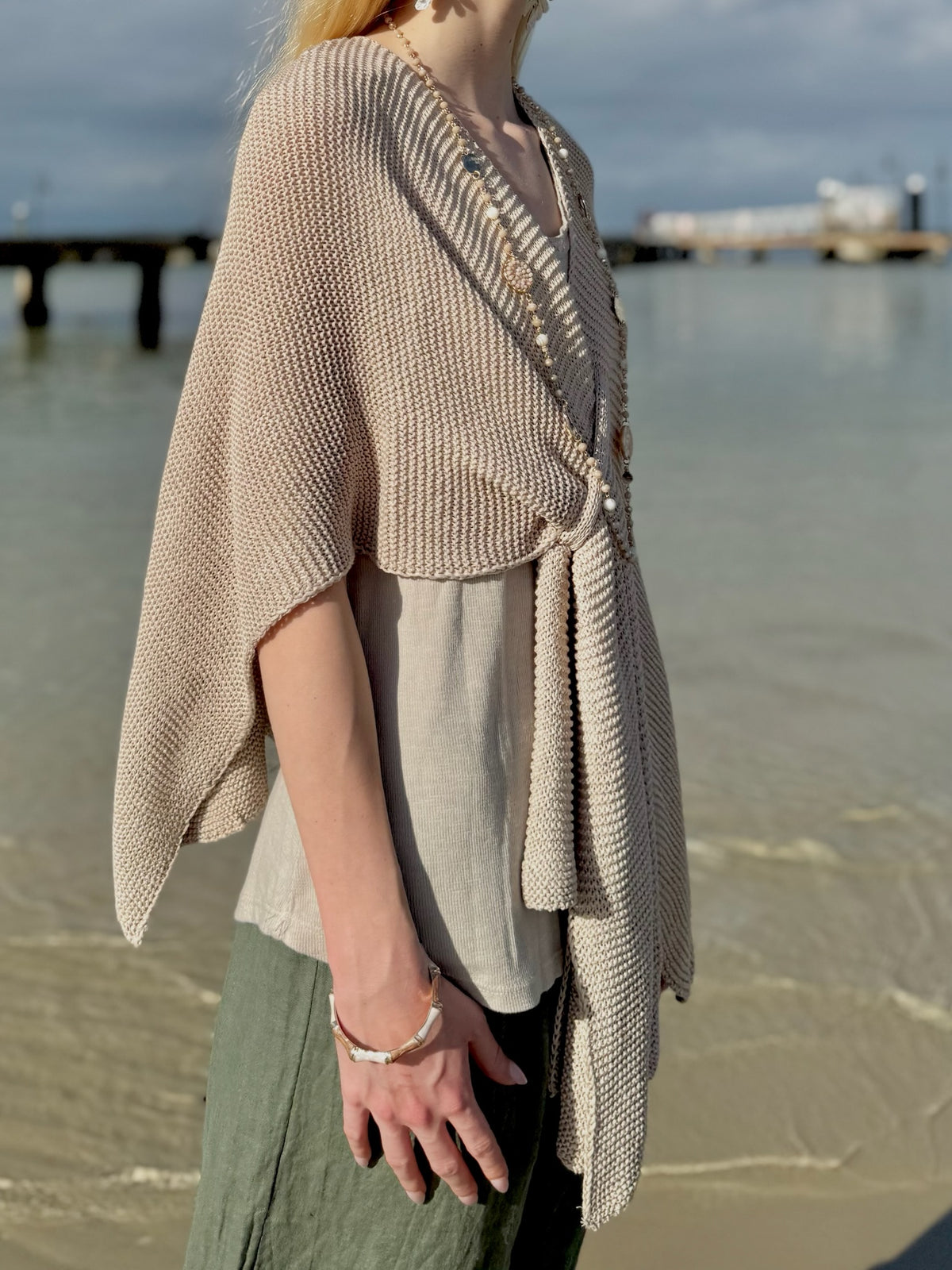 Side view of the Elise Wrap Poncho in Beige showing its textured knit, draped sleeves, and front tie detail, layered over a neutral tank top at Ardor Boutique.