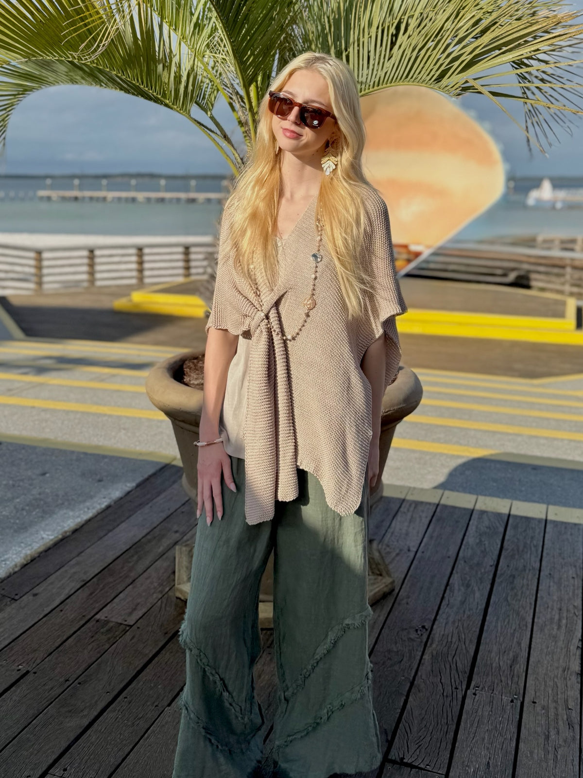 Model standing on a wooden boardwalk wearing a beige knit wrap poncho and olive wide-leg linen pants, with palm fronds and the ocean in the background.