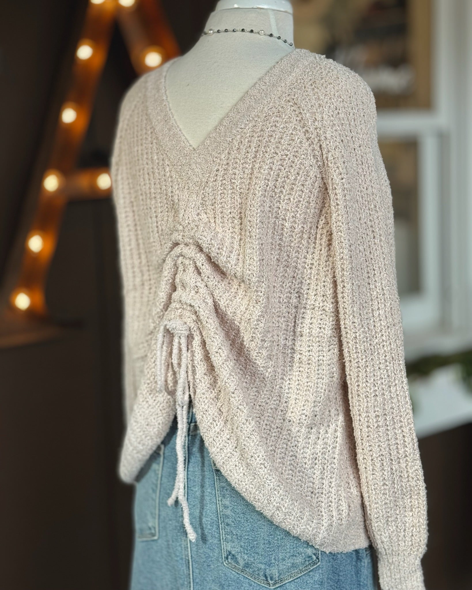 Side and back view of reversible neutral sweater with V neck and ruched drawstring worn at the back paired with denim skirt