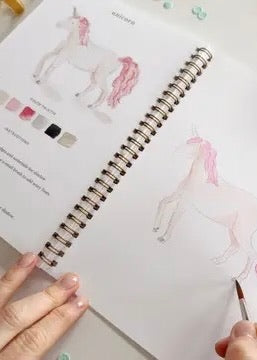 Open Fairytale watercolor workbook showing unicorn watercolor lesson with guided instructions and sketch ready to paint on thick paper