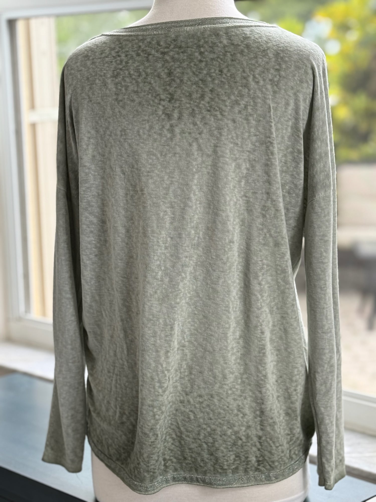 Back view of a sage green long-sleeve V-neck top with a casual, relaxed silhouette, shown on a mannequin by a window