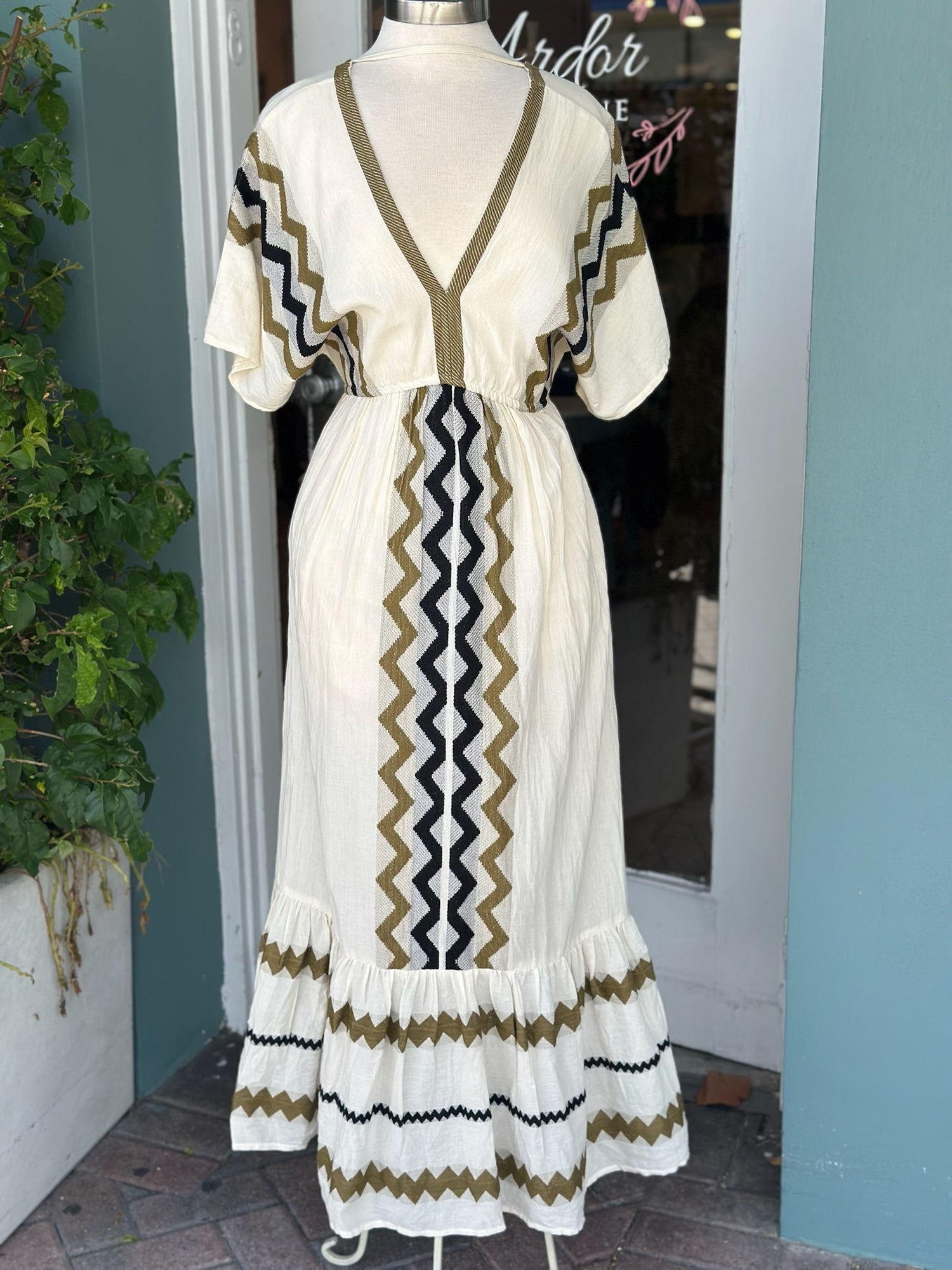 Cream-colored boho maxi dress displayed on a mannequin, featuring olive green, black, and white zigzag embroidery, short flutter sleeves, a V-neckline, and a tiered hem.