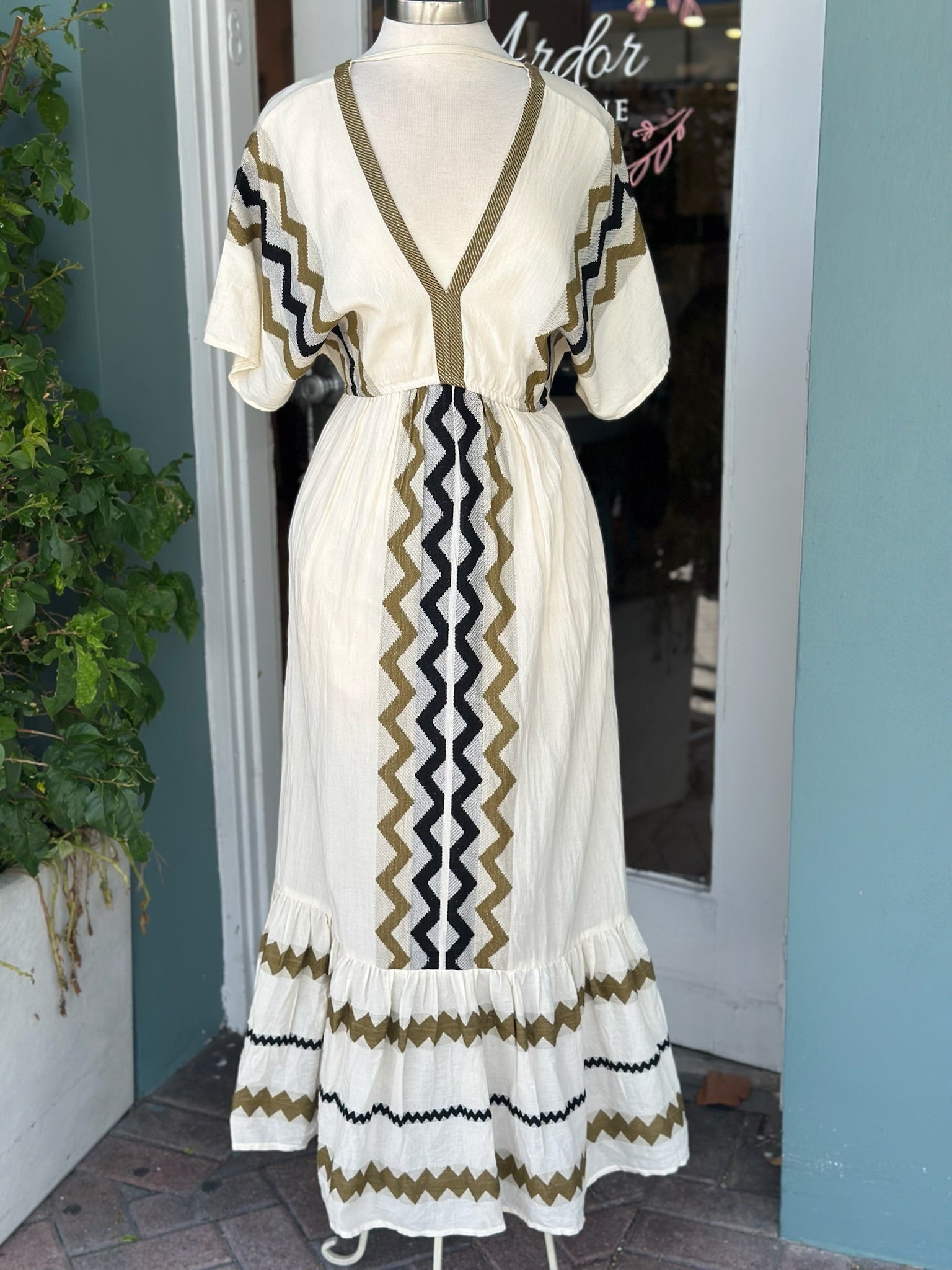 Cream-colored boho maxi dress displayed on a mannequin, featuring olive green, black, and white zigzag embroidery, short flutter sleeves, a V-neckline, and a tiered hem.