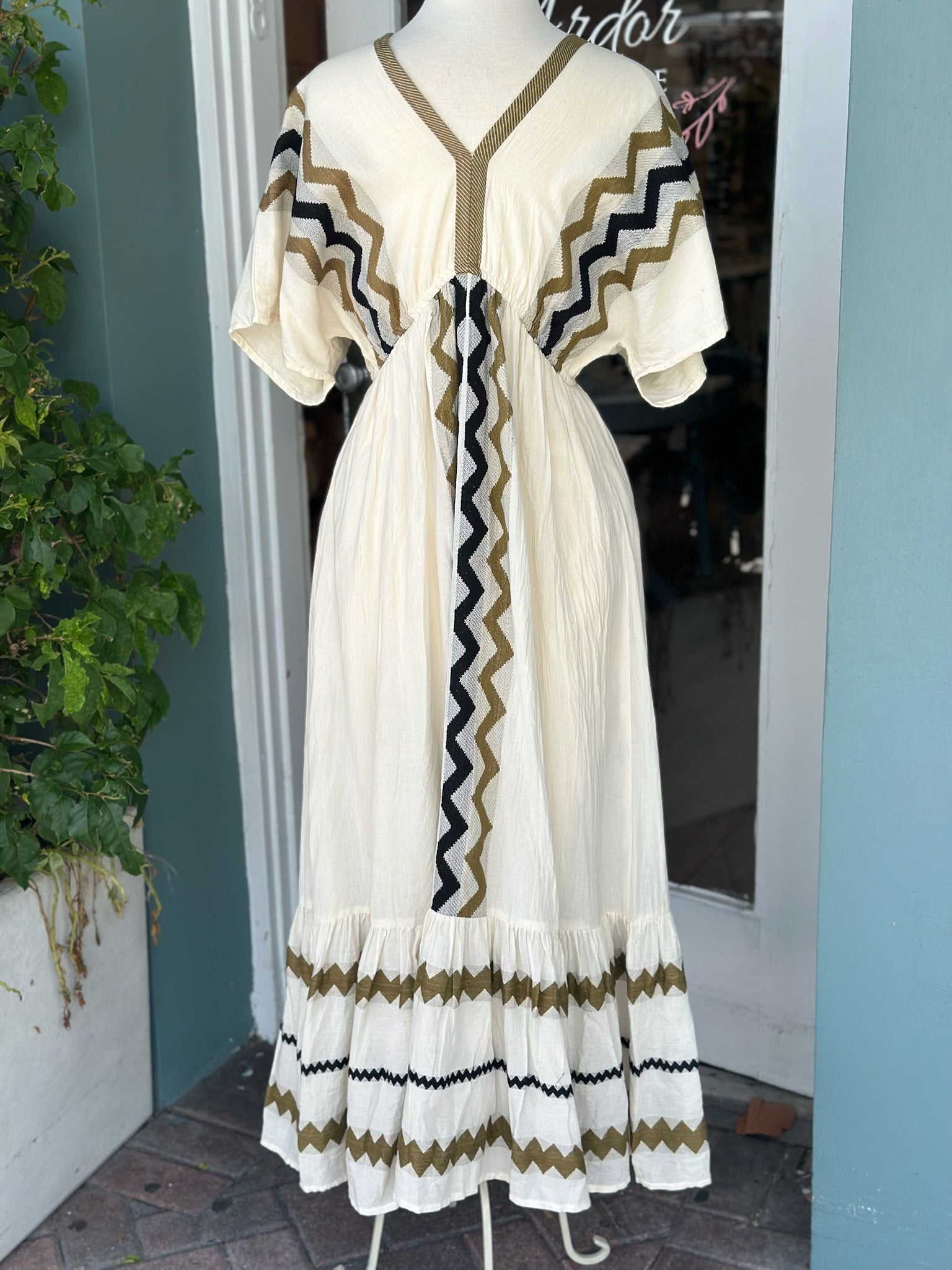Cream-colored boho maxi dress displayed on a mannequin, featuring olive green, black, and white zigzag embroidery, short flutter sleeves, a V-neckline, and a tiered hem.