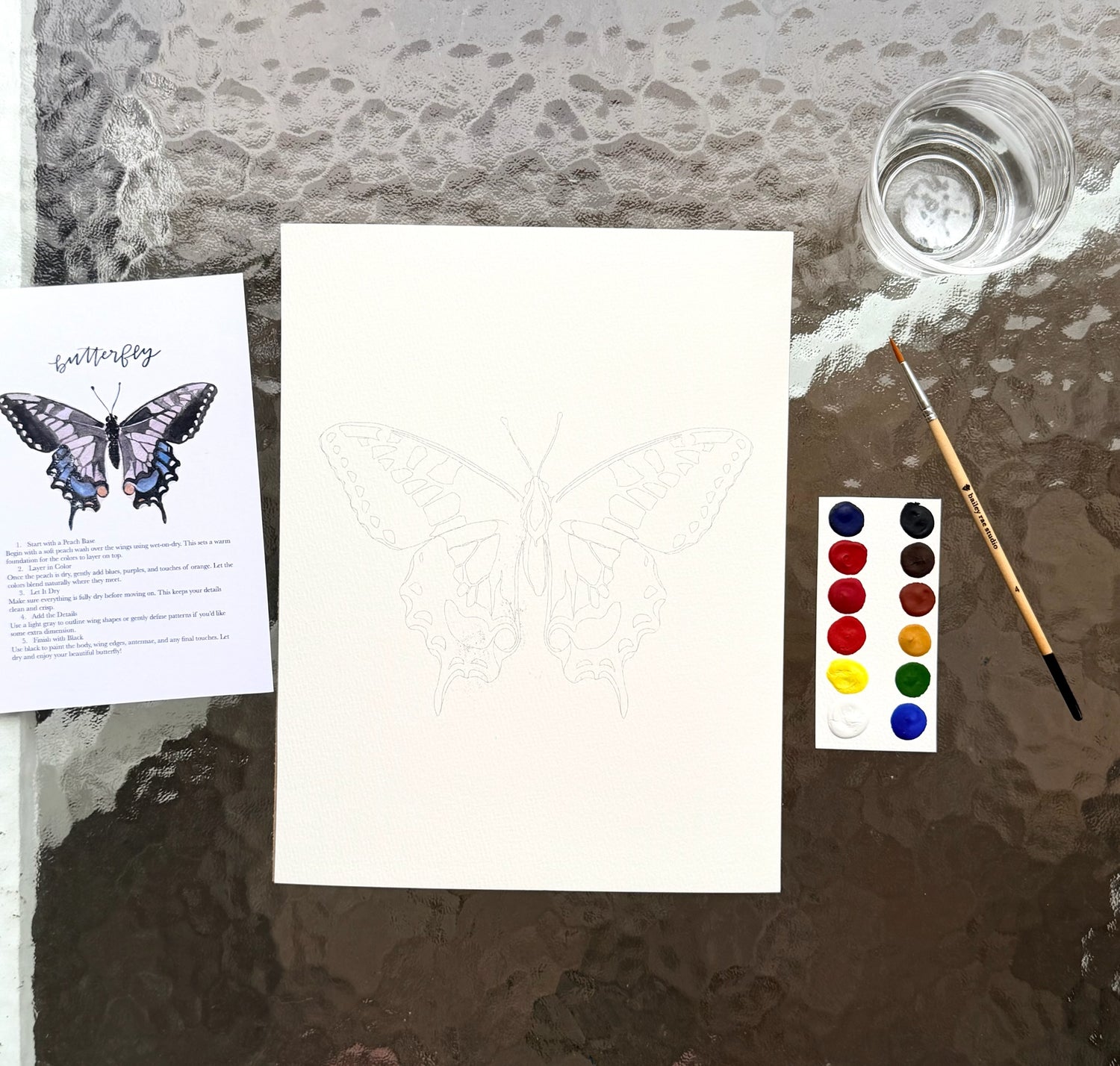 “Butterfly watercolor painting kit setup with butterfly outline on watercolor paper, instruction card, paint palette, brush, and cup of water on a glass table.”
