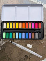 “Watercolor paint set with a range of bright and neutral colors, a refillable water brush, and sponge on a glass table.”