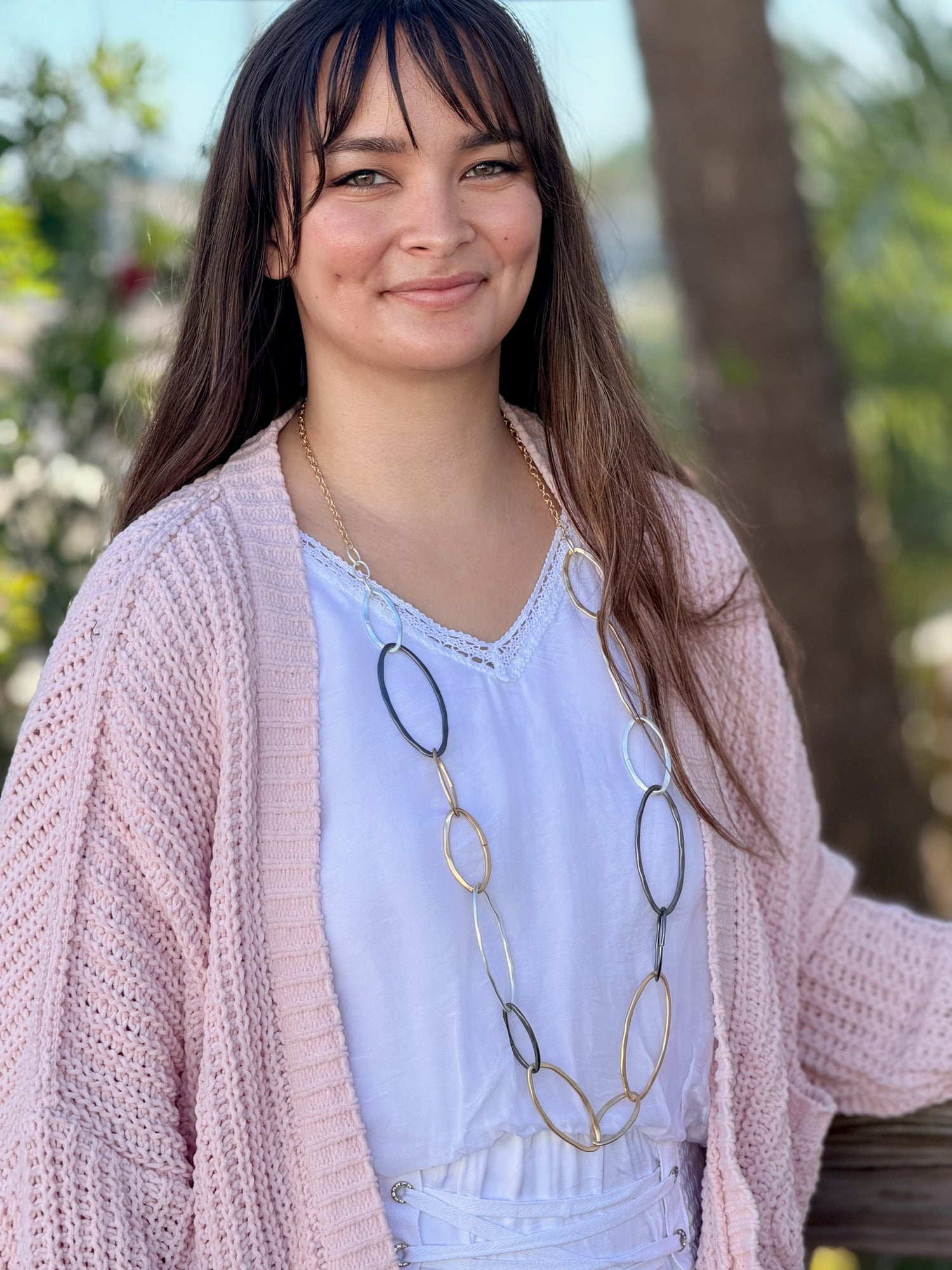 Woman wearing a pink cardigan and white blouse with a long necklace outdoors.