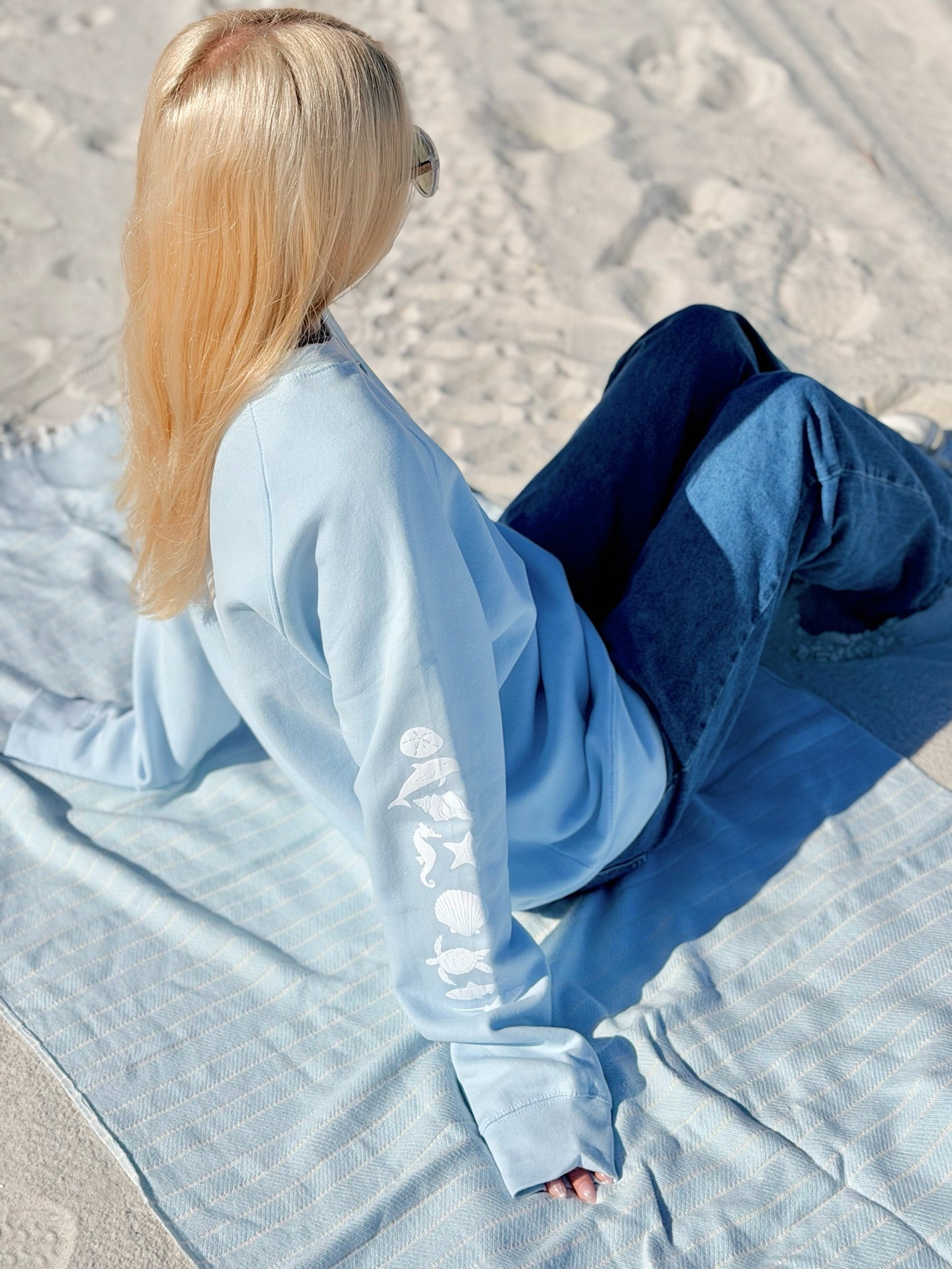 Person sitting on a blanket in a sandy area wearing a light blue outfit.