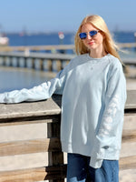 Person wearing a light blue sweatshirt with sea life icons on sleeve by a waterfront