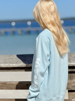 Person wearing a light blue sweatshirt with a graphic design by the water