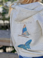 Oatmeal hoodie with butterfly design and text on the back, worn by a person.