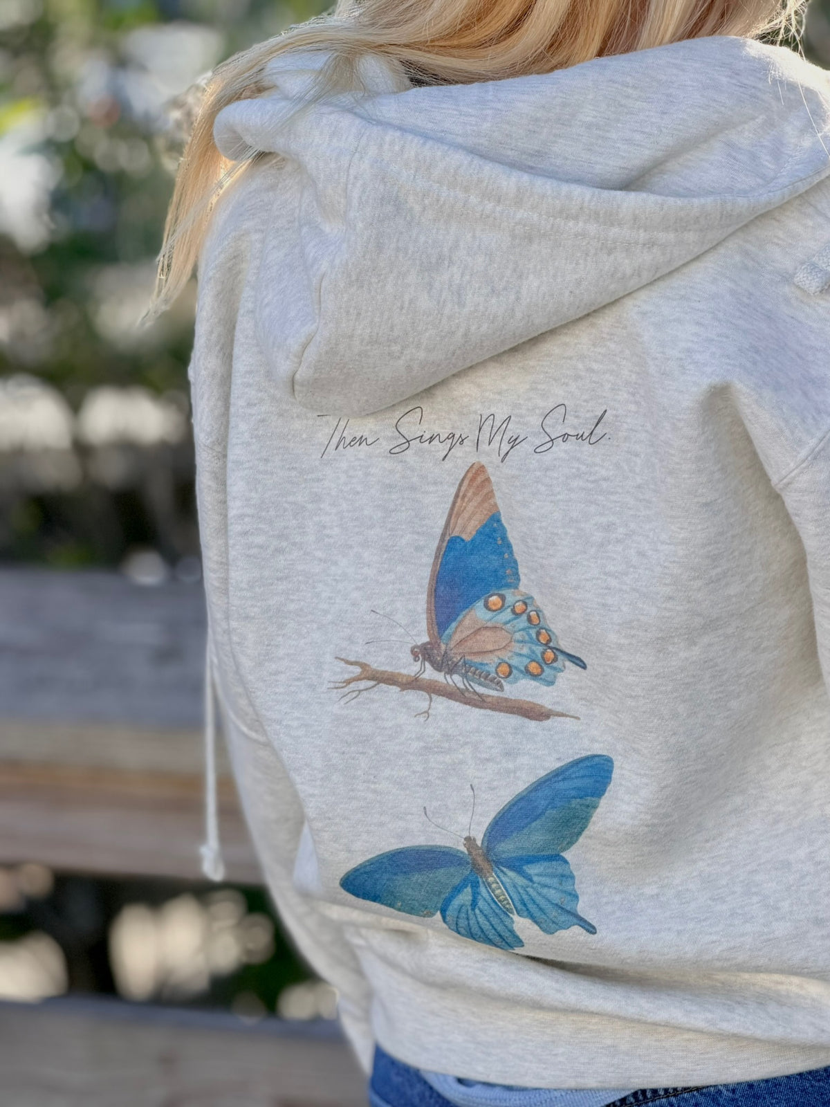 Oatmeal hoodie with butterfly design and text on the back, worn by a person.