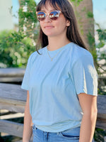 Person wearing a Sea Mist T-Shirt and sunglasses outdoors with greenery in the background