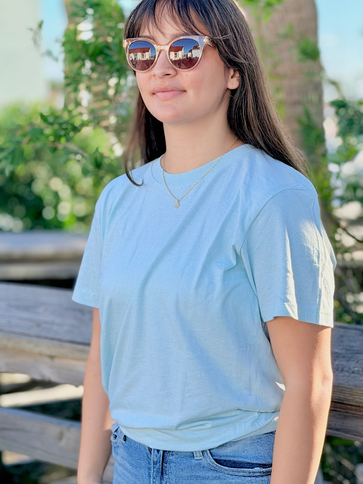 Person wearing a Sea Mist T-Shirt and sunglasses outdoors with greenery in the background