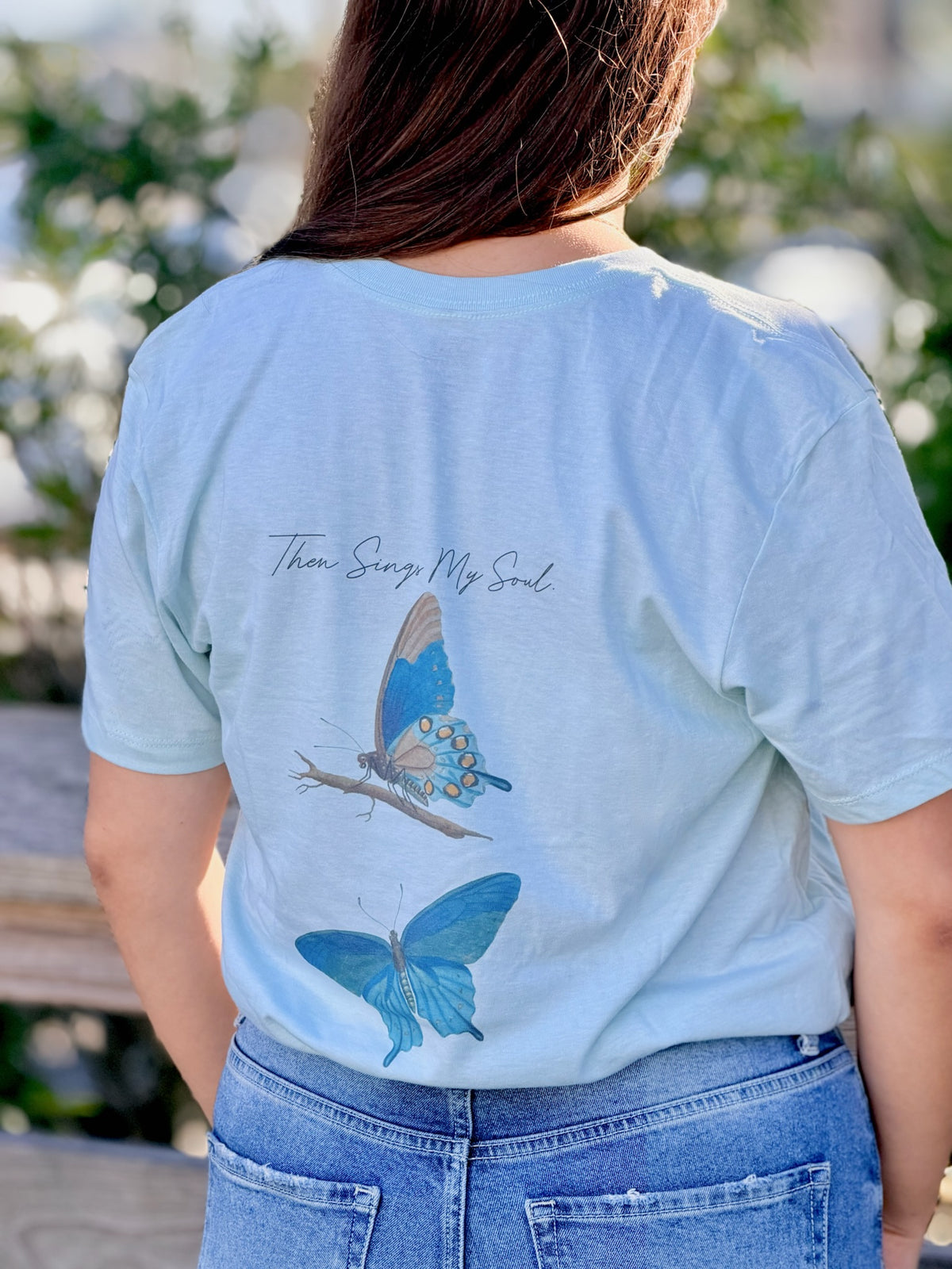 Sea Mist t-shirt with butterfly design and text on the back, worn by a person outdoors.