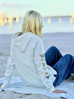 Person sitting on a beach wearing a white hoodie with butterfly designs and blue jeans.