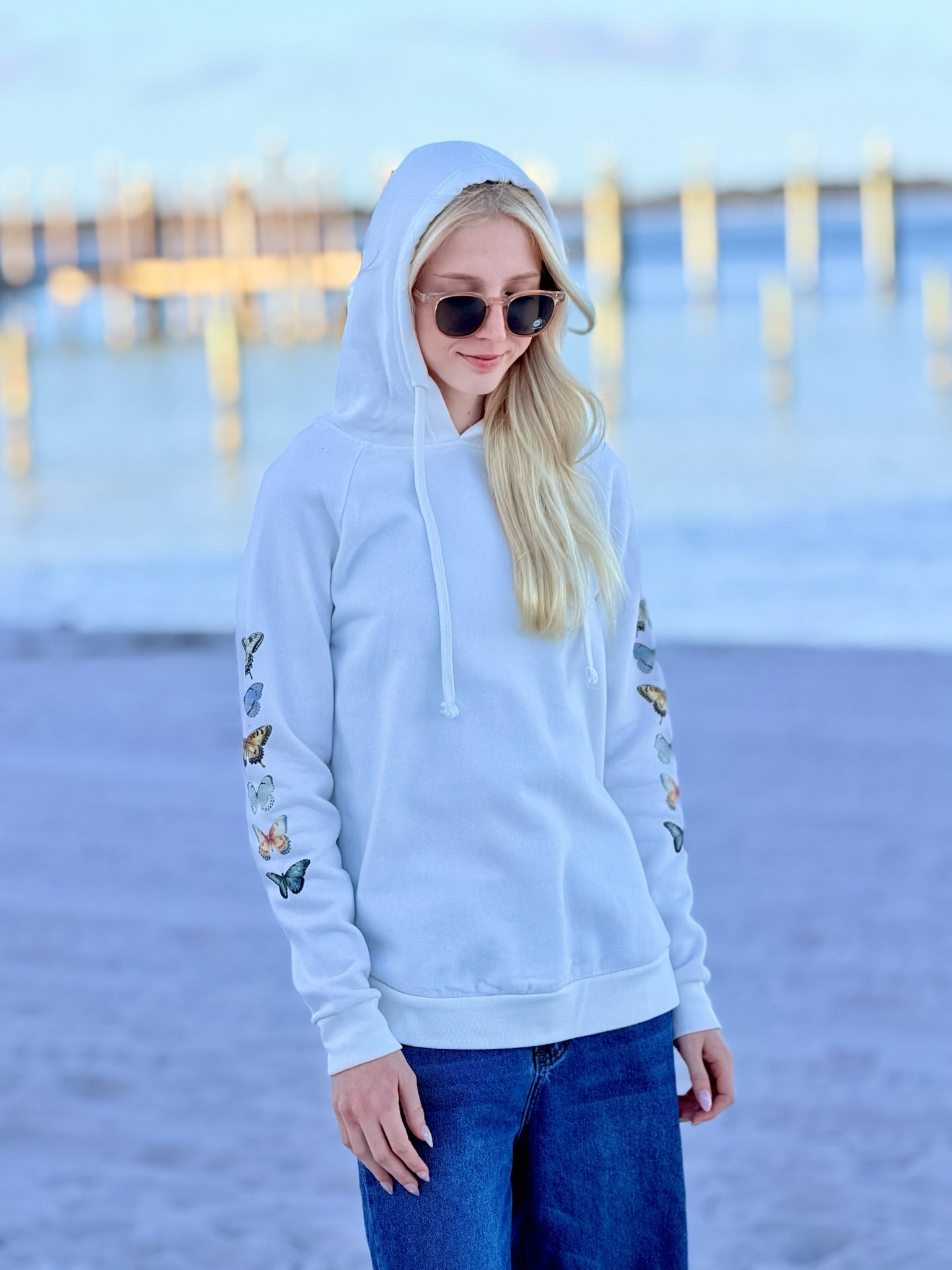 Woman wearing a white hoodie with butterfly designs on the sleeves at the beach.