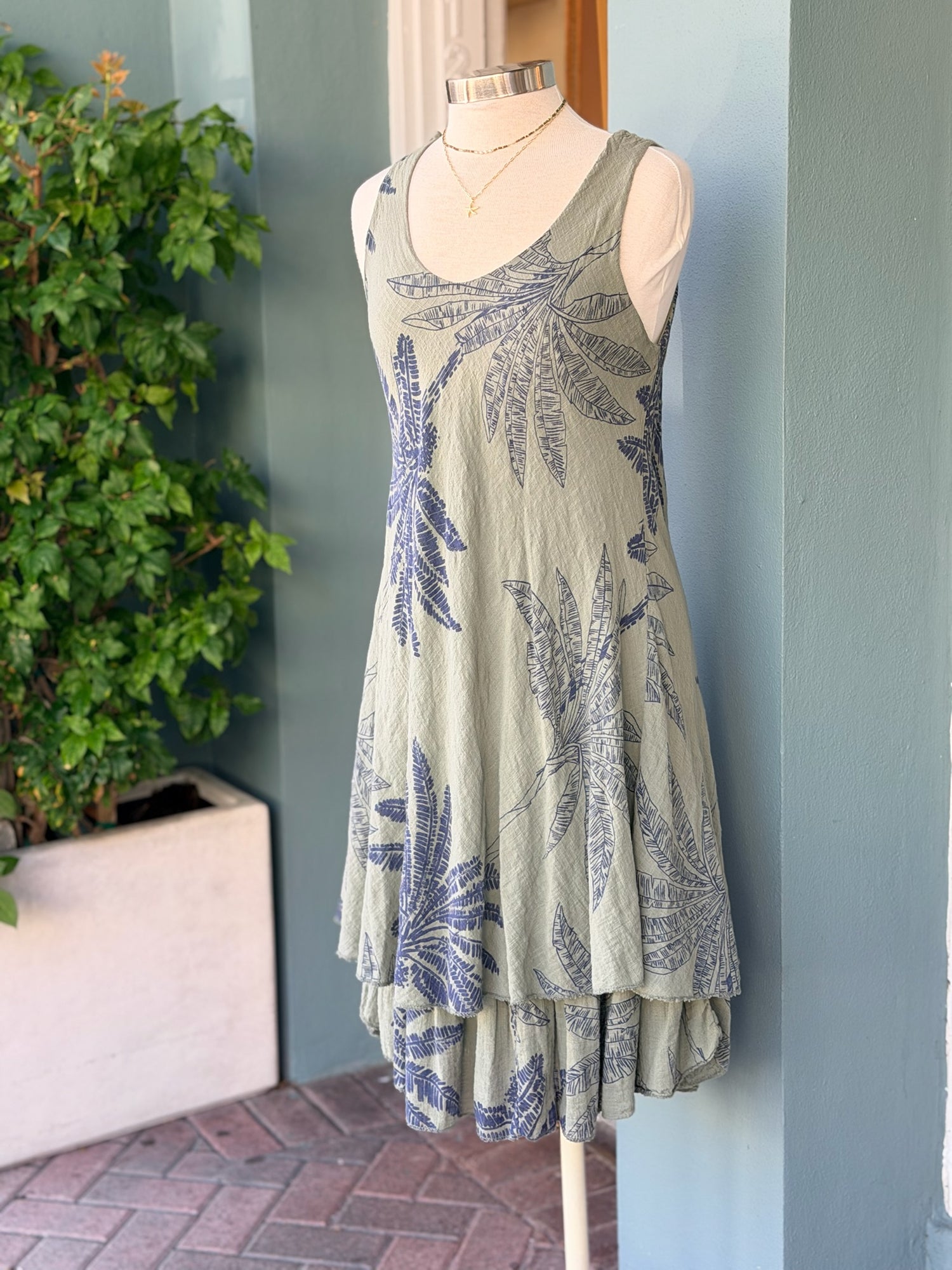 Sleeveless olive dress with navy palm print and a flowy layered hem, displayed on a mannequin.