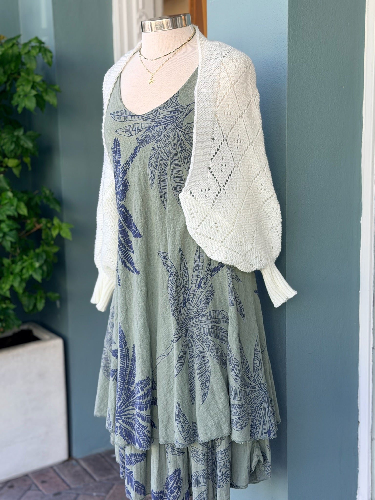 Sleeveless olive palm print dress in navy tones layered with a white open-knit cardigan, displayed on a mannequin.