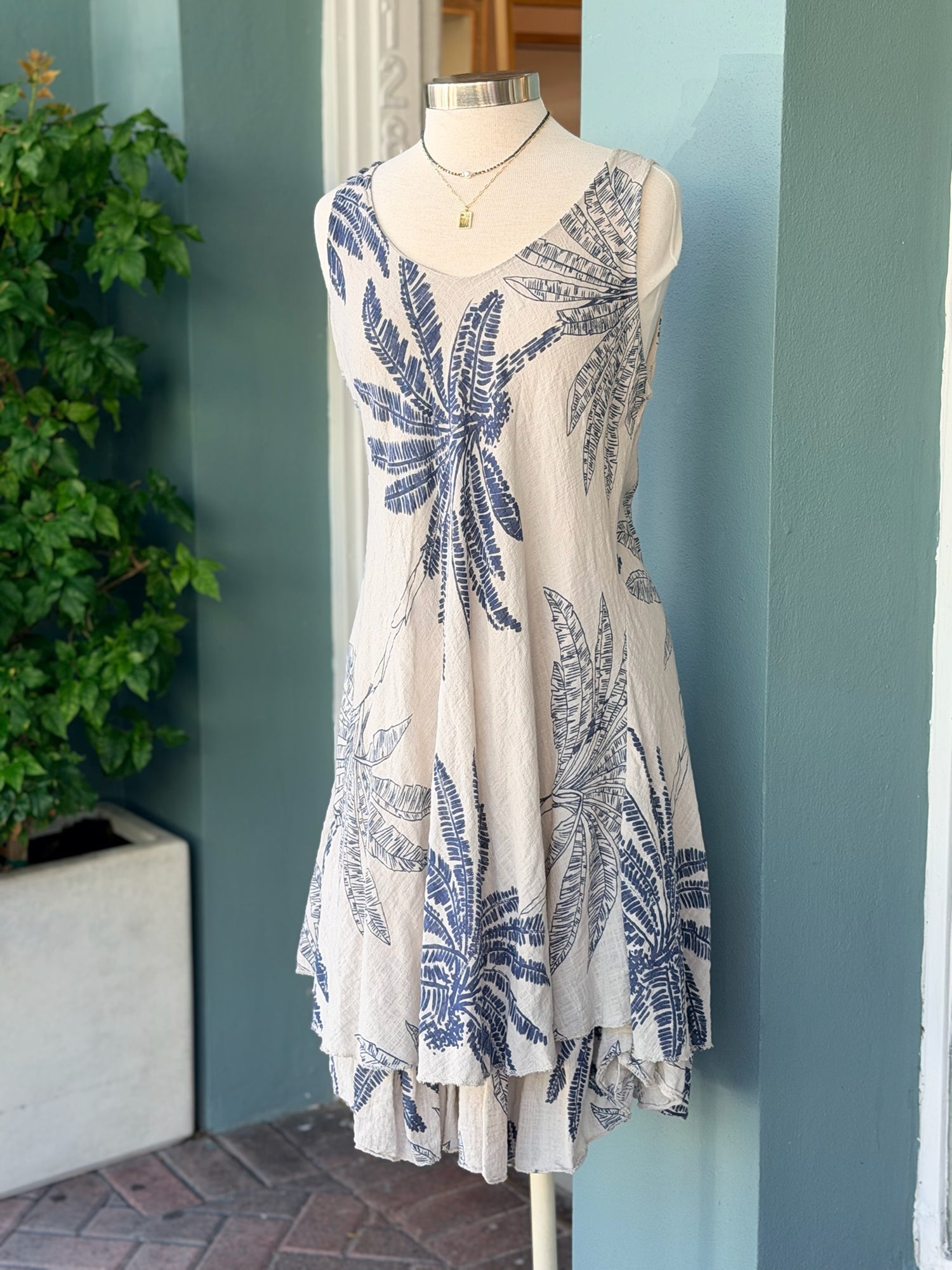 Sleeveless taupe dress with navy palm print, featuring a flowy layered hem and lightweight fabric, displayed on a mannequin.