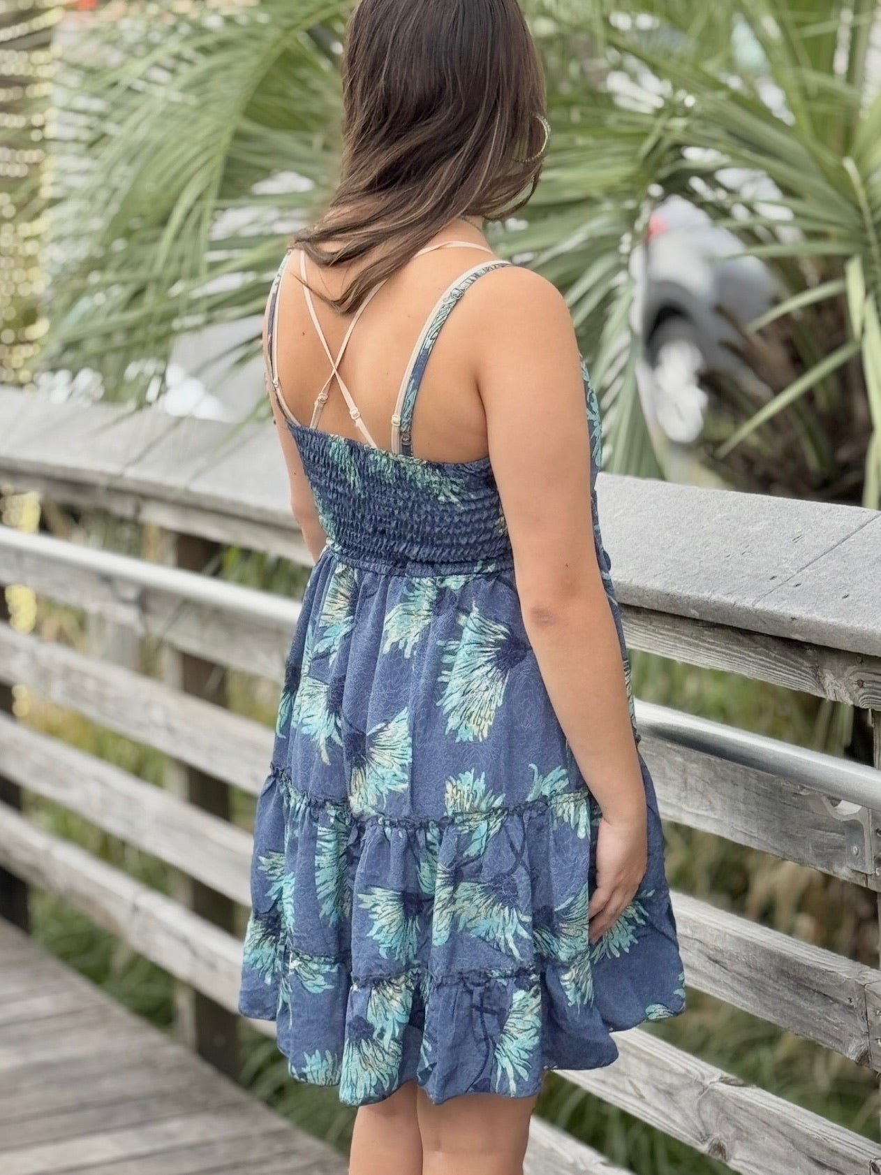 Model wearing a short blue dress with aqua floral print, styled with gold jewelry and sunglasses back view