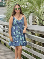Model wearing a short blue dress with aqua floral print, styled with gold jewelry and sunglasses
