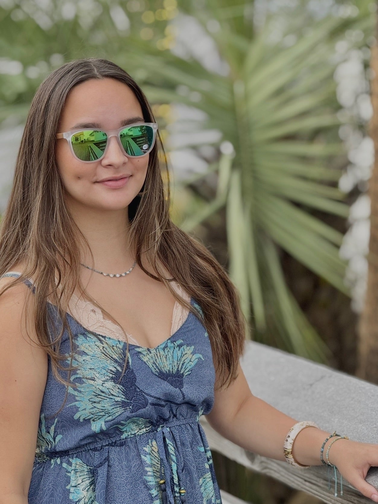 Model wearing a short blue dress with aqua floral print, styled with gold jewelry and sunglasses
