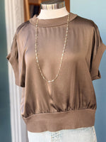 Taupe short-sleeve blouse on a mannequin with a necklace against a light blue wall.