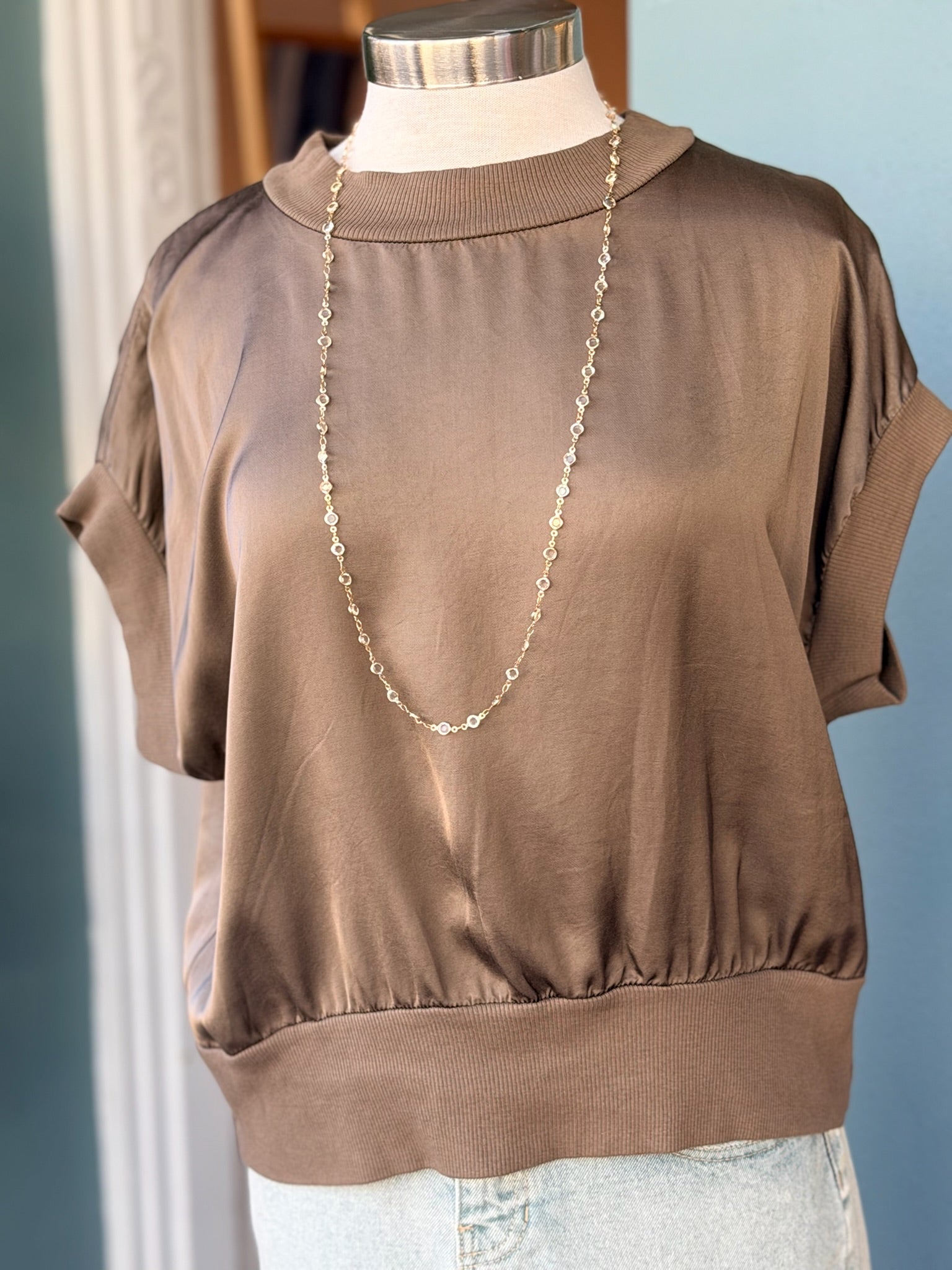Taupe short-sleeve blouse on a mannequin with a necklace against a light blue wall.