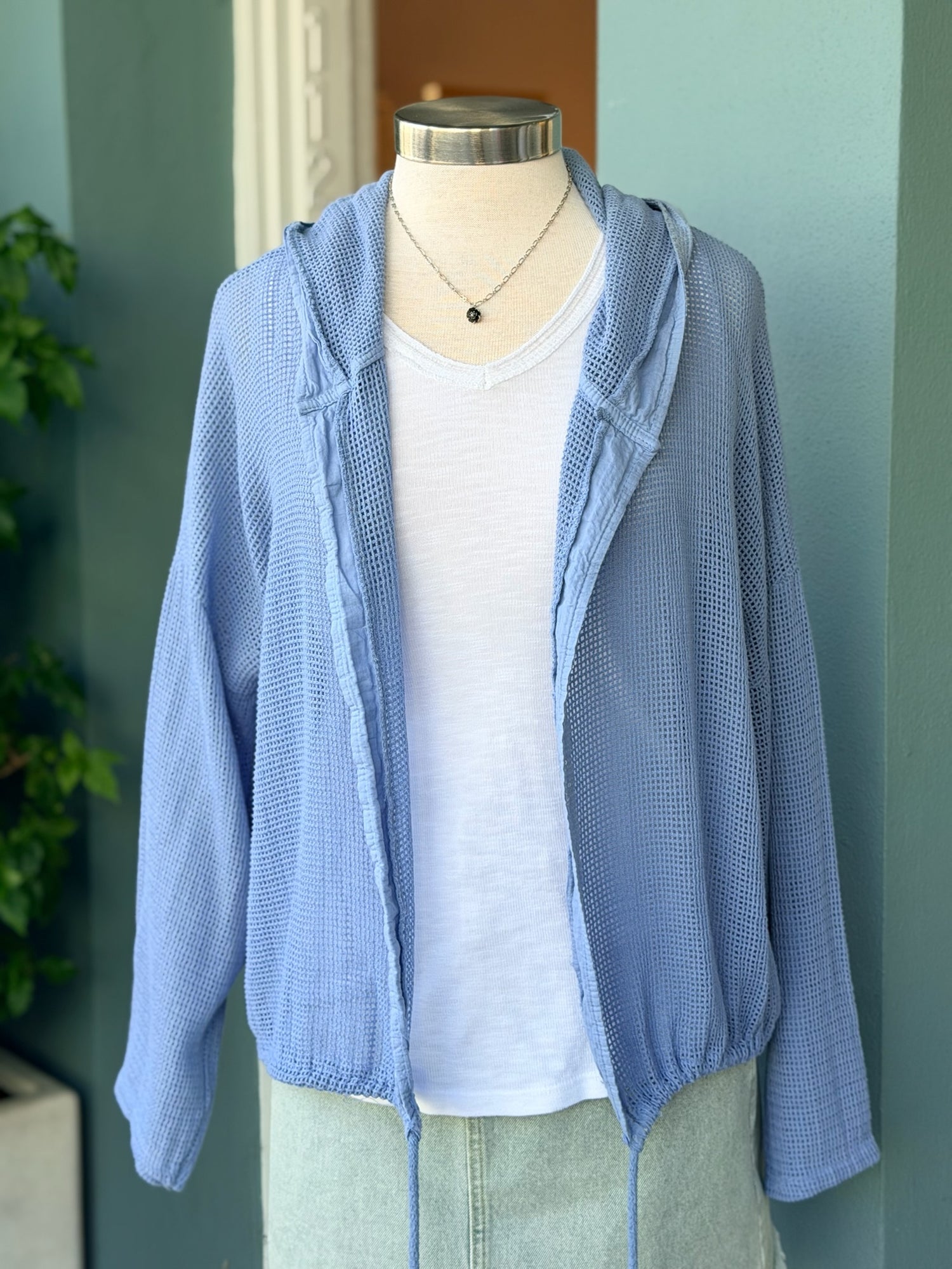 Mannequin wearing a light blue cardigan over a white shirt against a green wall.