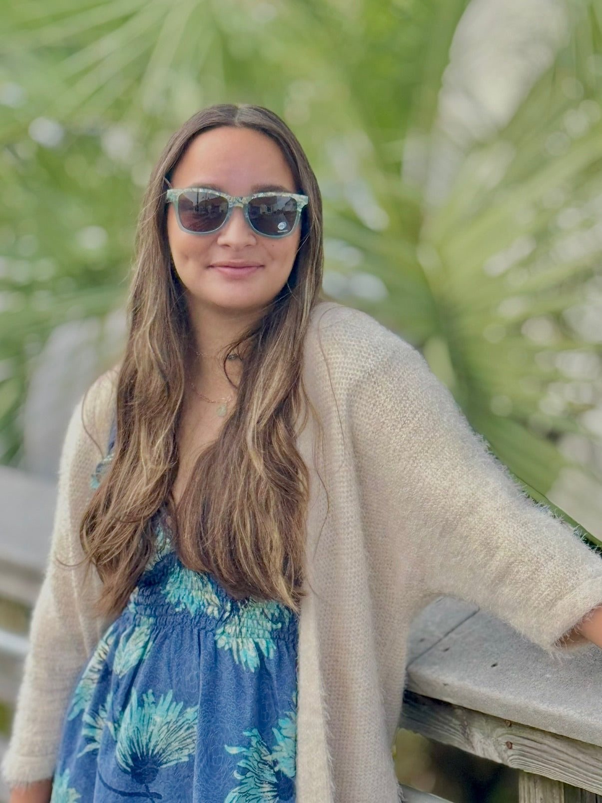 Woman wearing sunglasses and a beige cardigan over a blue dress with a green leafy background
