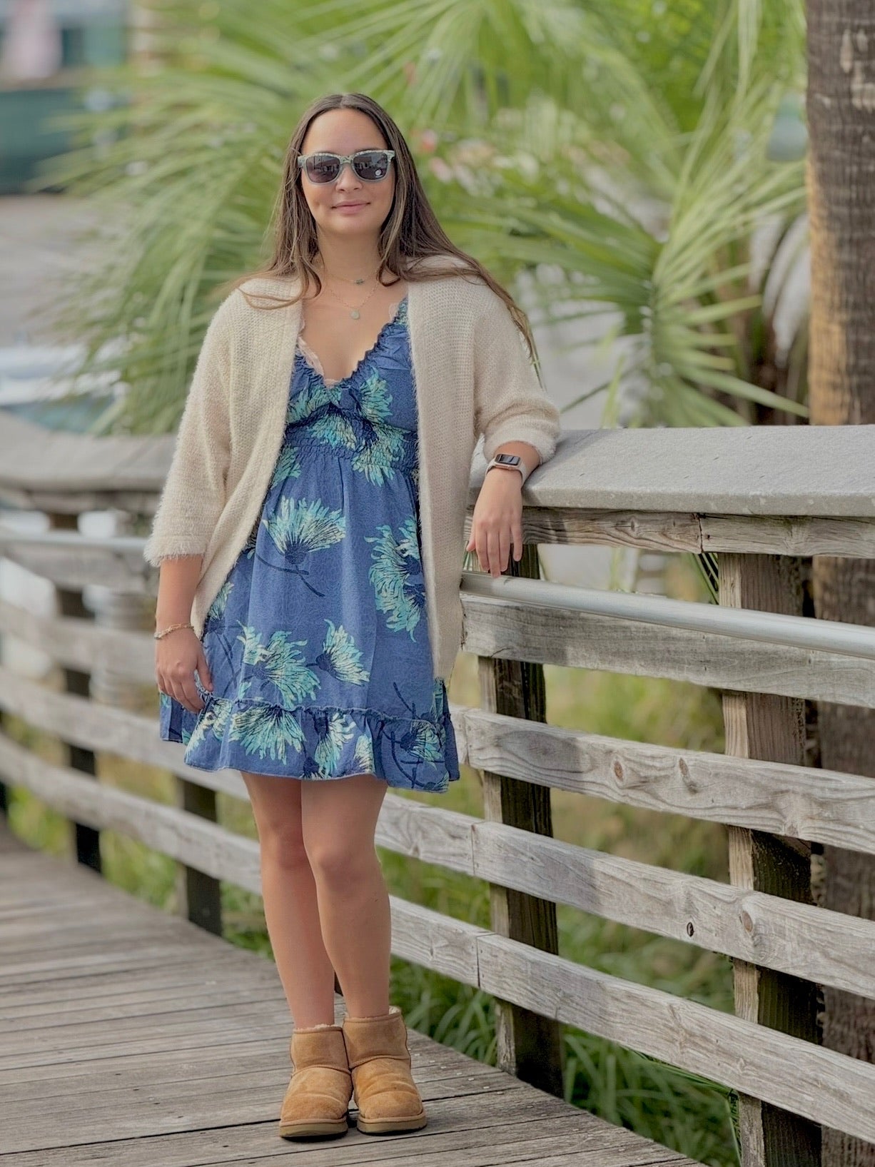 Woman in a blue floral dress and beige cardigan standing on a wooden boardwalk with palm trees in the background.