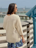 Woman in a beige sweater and floral dress standing on a wooden pier by the water.