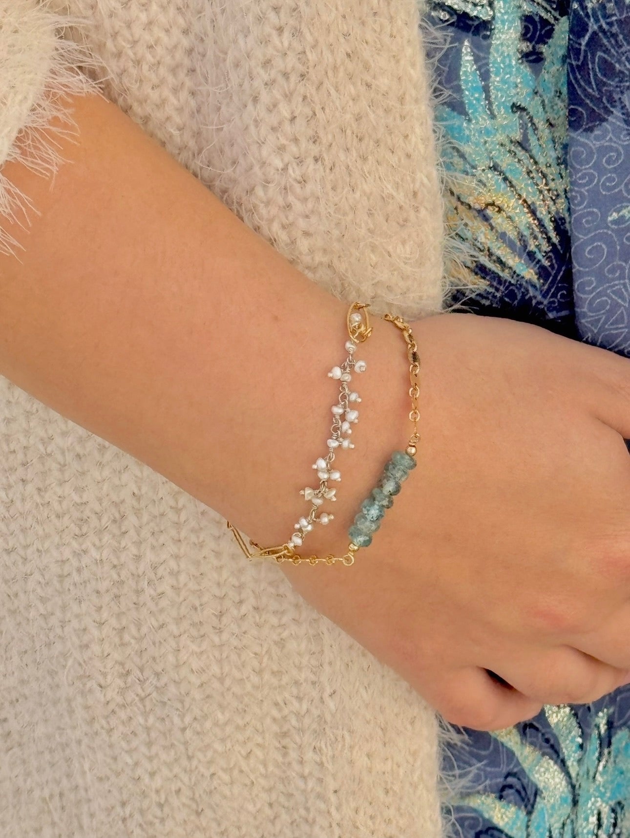 Close-up of a hand wearing two gold bracelets with blue beads on a textured surface.