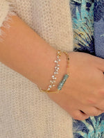 Close-up of a hand wearing two gold bracelets with blue beads on a textured surface.