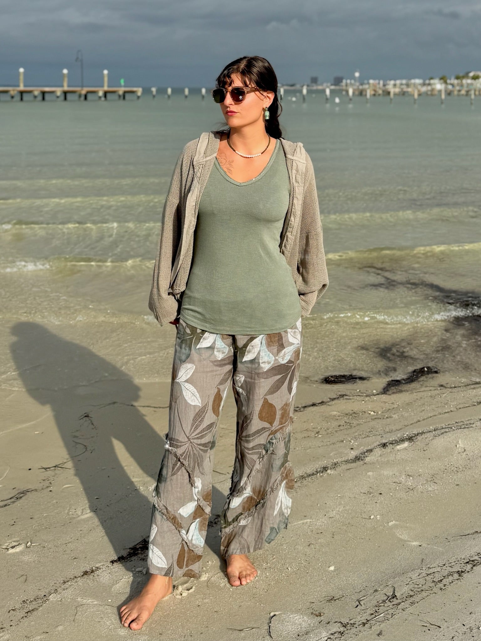 Model standing barefoot on the beach wearing the Catherine Linen Pants in mocha floral with an olive tank and taupe mesh jacket, waves in the background.