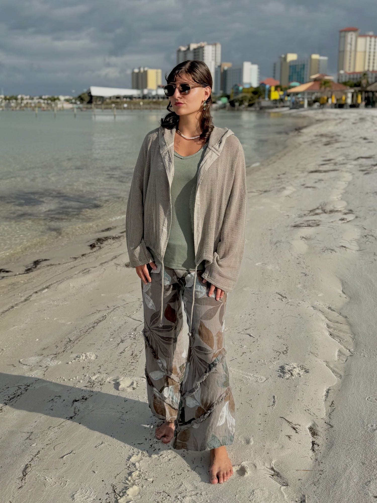 Model standing on the beach wearing the taupe Gia Mesh Hooded Jacket over a sage tank and patterned pants, with the shoreline and city buildings in the background.