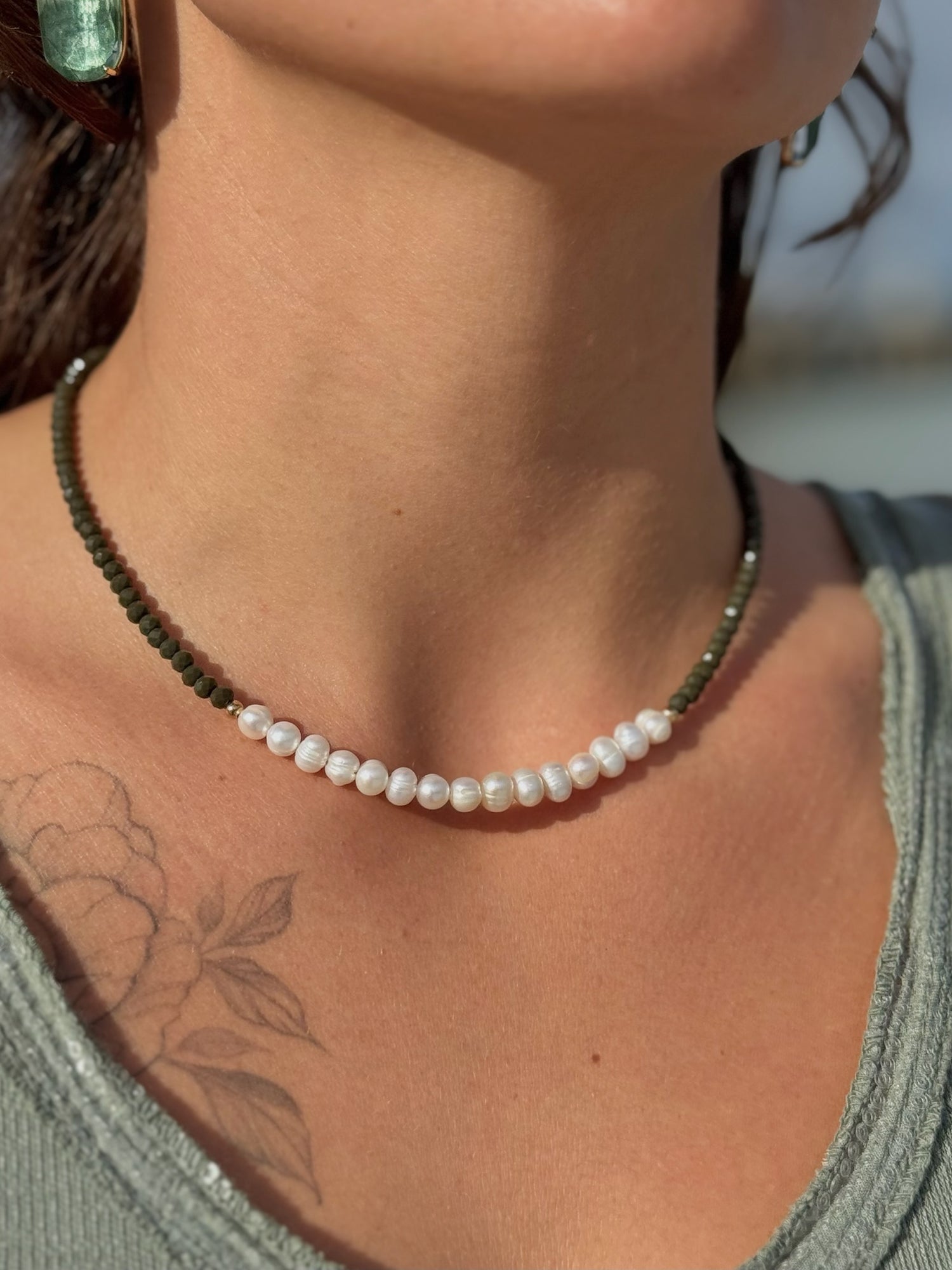 A woman wearing the Hosanna necklace in olive, showing deep olive beads and freshwater pearls resting at the collarbone in natural sunlight.