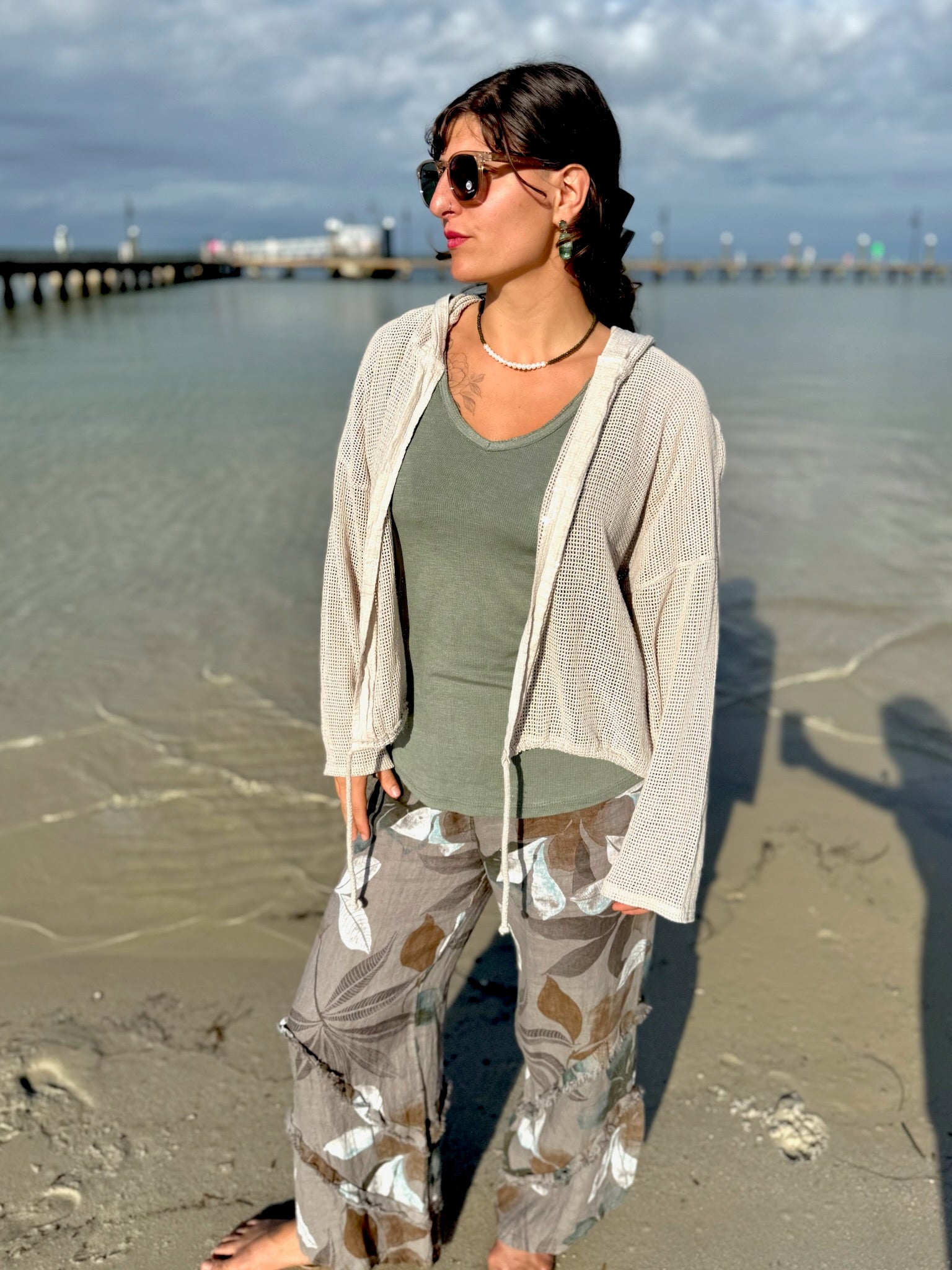 Model standing on the shoreline wearing the beige Gia Mesh Hooded Jacket over a sage green tank and patterned wide-leg pants, with soft sunlight highlighting the mesh texture.