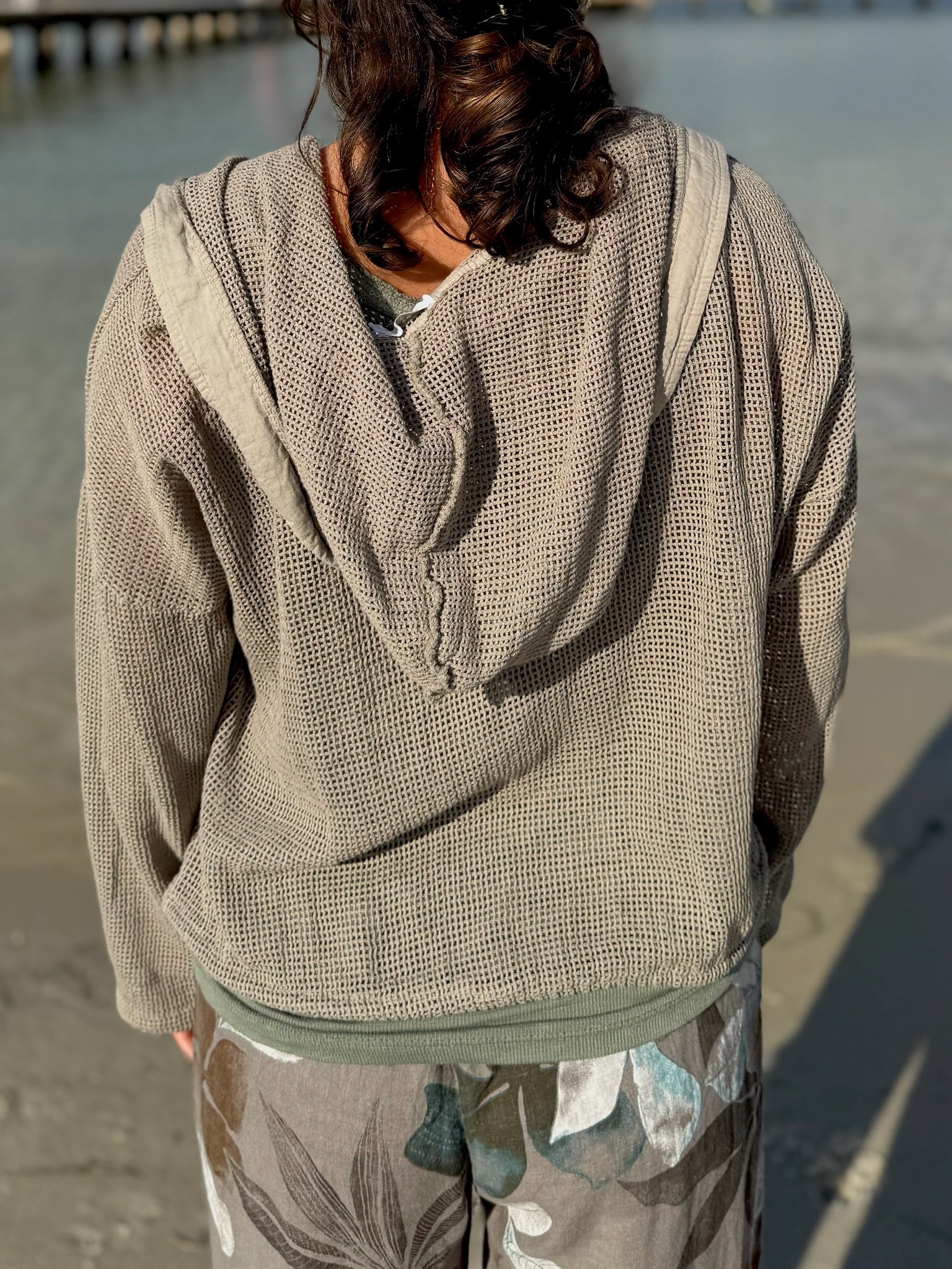 Back view of the taupe Gia Mesh Hooded Jacket showing the open-knit texture, relaxed hood, and loose fit in sunlight by the water.