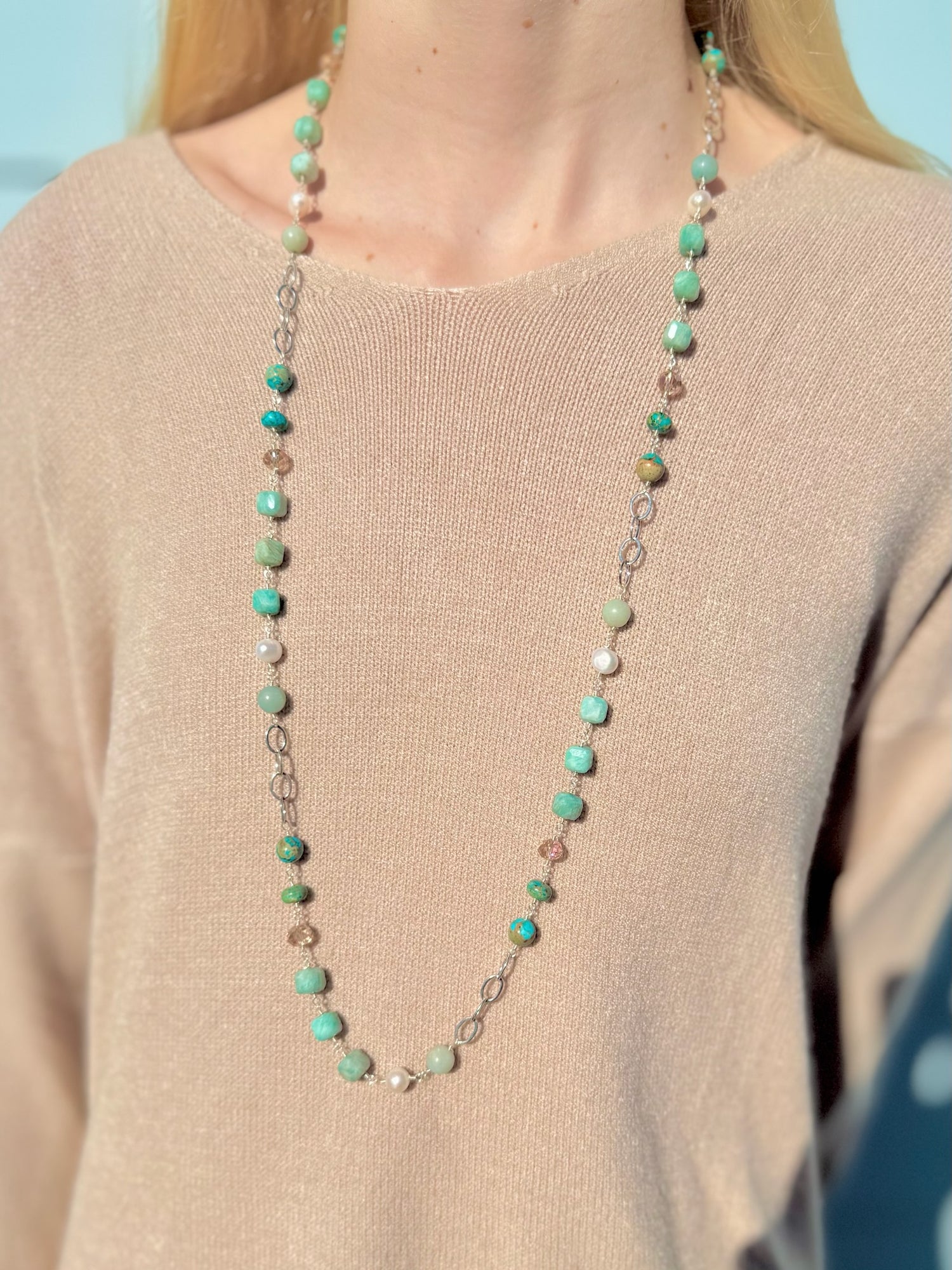 Long necklace with green beads and silver chain on a beige sweater against a blue background