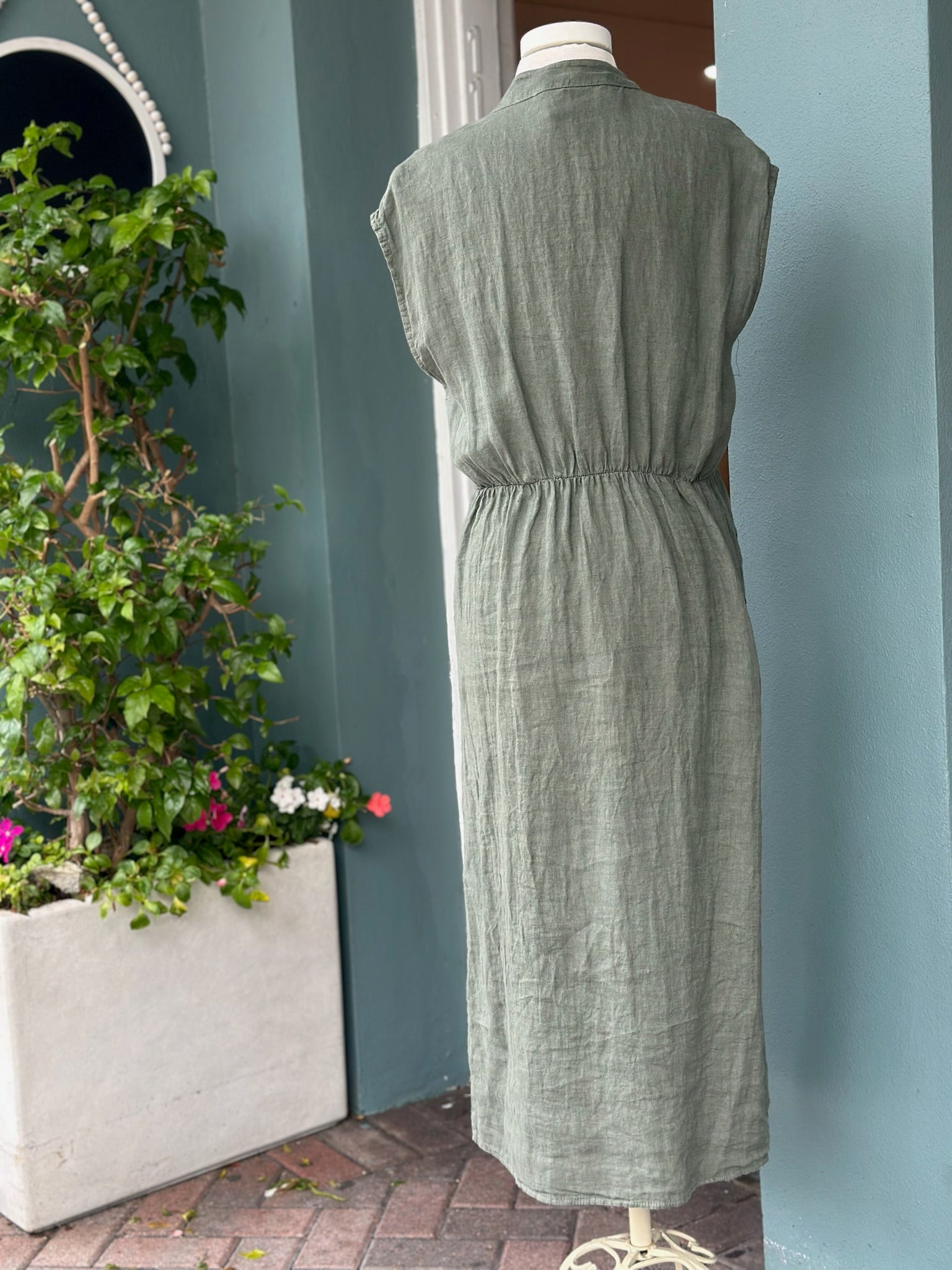 Back view of the Sharon Dress in rich olive green linen featuring cap sleeves, an elasticized waist detail, and a relaxed midi-length silhouette. Made from lightweight, breathable linen with a naturally textured finish.