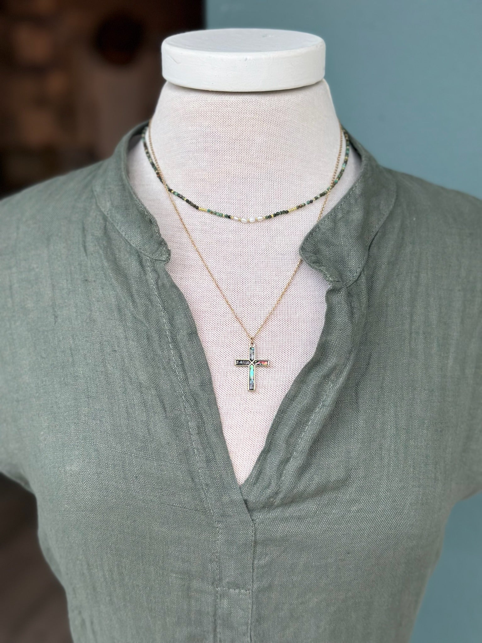 Abalone shell cross pendant necklace with iridescent blue and green tones, layered with green gemstone beads and styled with an olive green linen dress.
