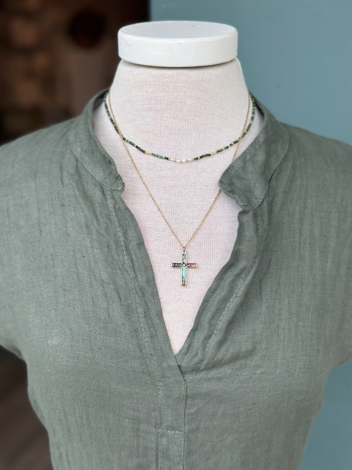 Close-up of the Sharon Dress in rich olive green linen, showcasing the split neckline, soft crinkle texture, and delicate beaded necklace with a cross pendant. The breathable linen fabric and natural olive tone create an understated, coastal-inspired look.
