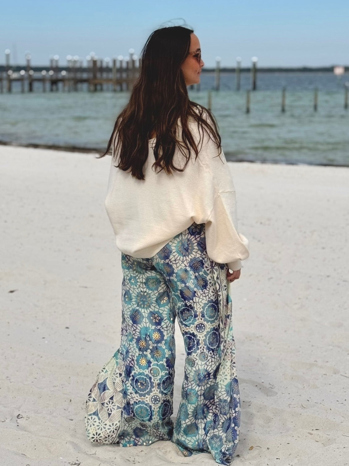 Woman wearing a white blouse and floral pants standing on a sandy beach with water and dock in the background.