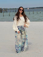 Woman in a white sweater and floral flowy pants standing on a sandy beach with water and dock in the background.